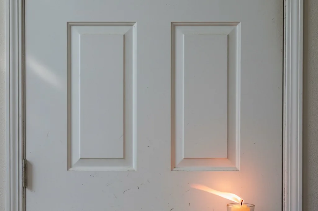 A lit candle flame bending sideways near the bottom corner of a closed white front door